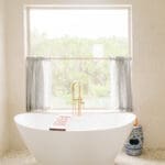 Freestanding tub and brass filler beneath a curtained window in a bathroom remodel