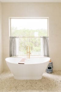 Freestanding tub and brass filler beneath a curtained window in a bathroom remodel
